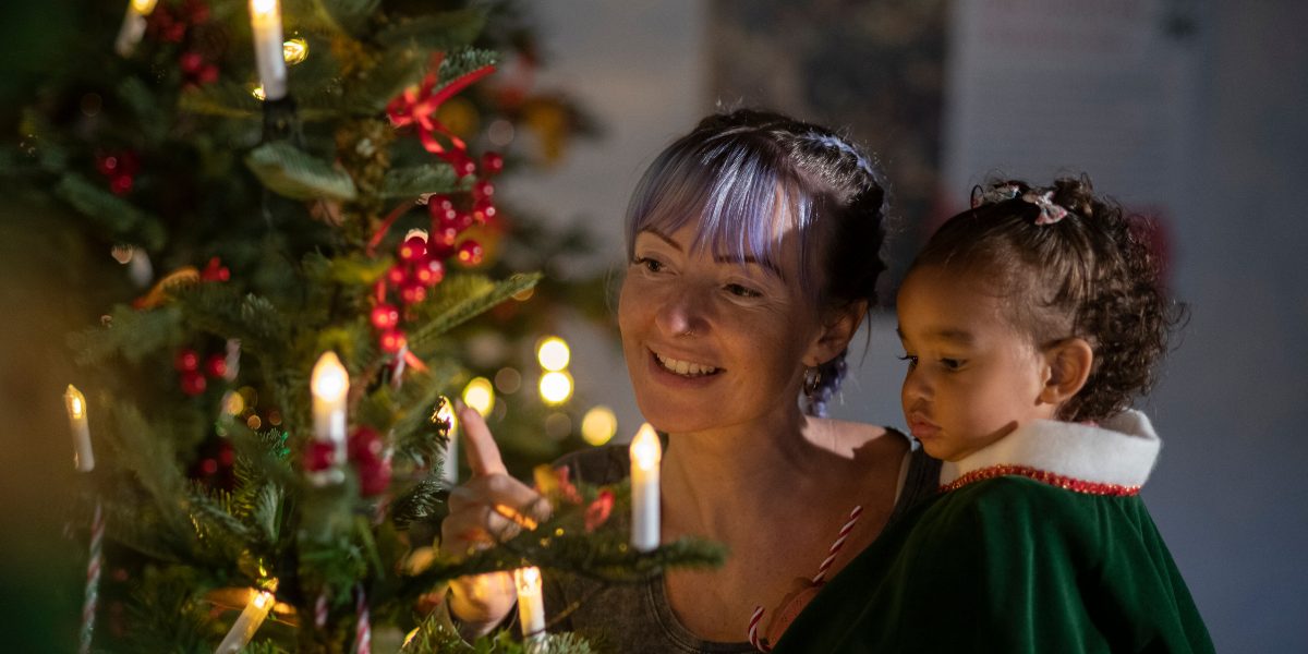 Adult carrying child with green cloak looking at Christmas tree decorations